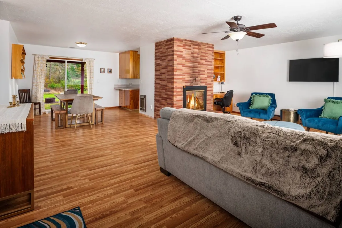 This image features a welcoming interior space centered around a cozy fireplace, a dining table with blue chairs, and a comfortable sofa. The room's arrangement encourages gatherings for meals, relaxation by the fire, and casual get-togethers. The décor blends rustic charm with modern touches, creating an inviting atmosphere.