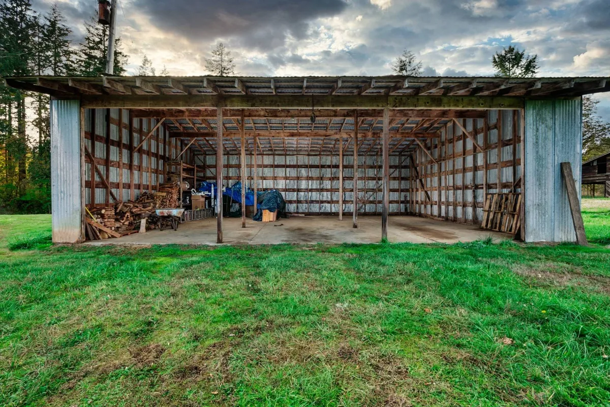 An open barn structure flanked by stacks of wood and covered by blue tarps, set against a rural backdrop. The rustic setting suggests a working farm environment and hints at the authentic, countryside character of the property.