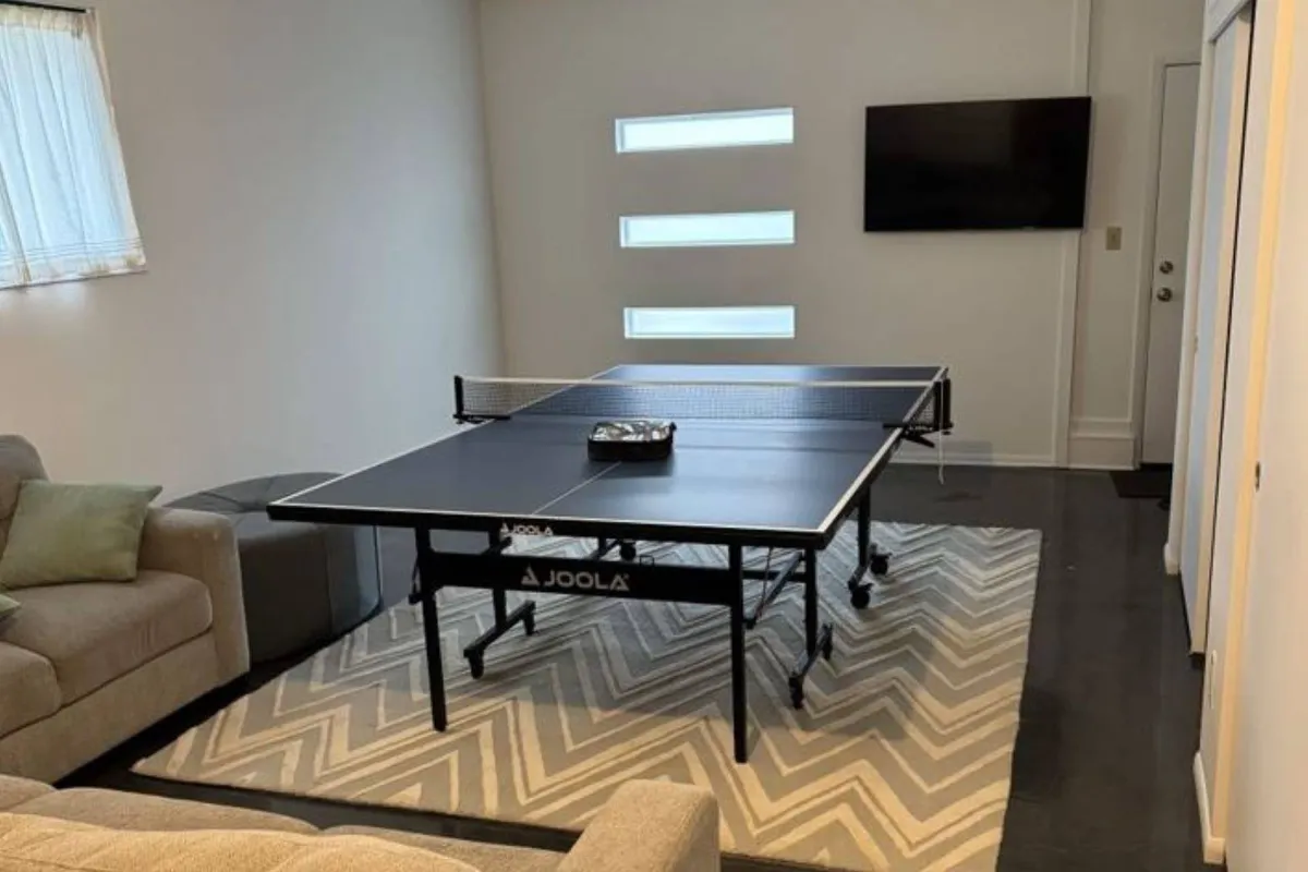 An energetic recreation room scene showing a ping pong table at center stage, complemented by a cozy sofa and a TV in the background. The layout highlights space for both friendly competition and post-game relaxation.