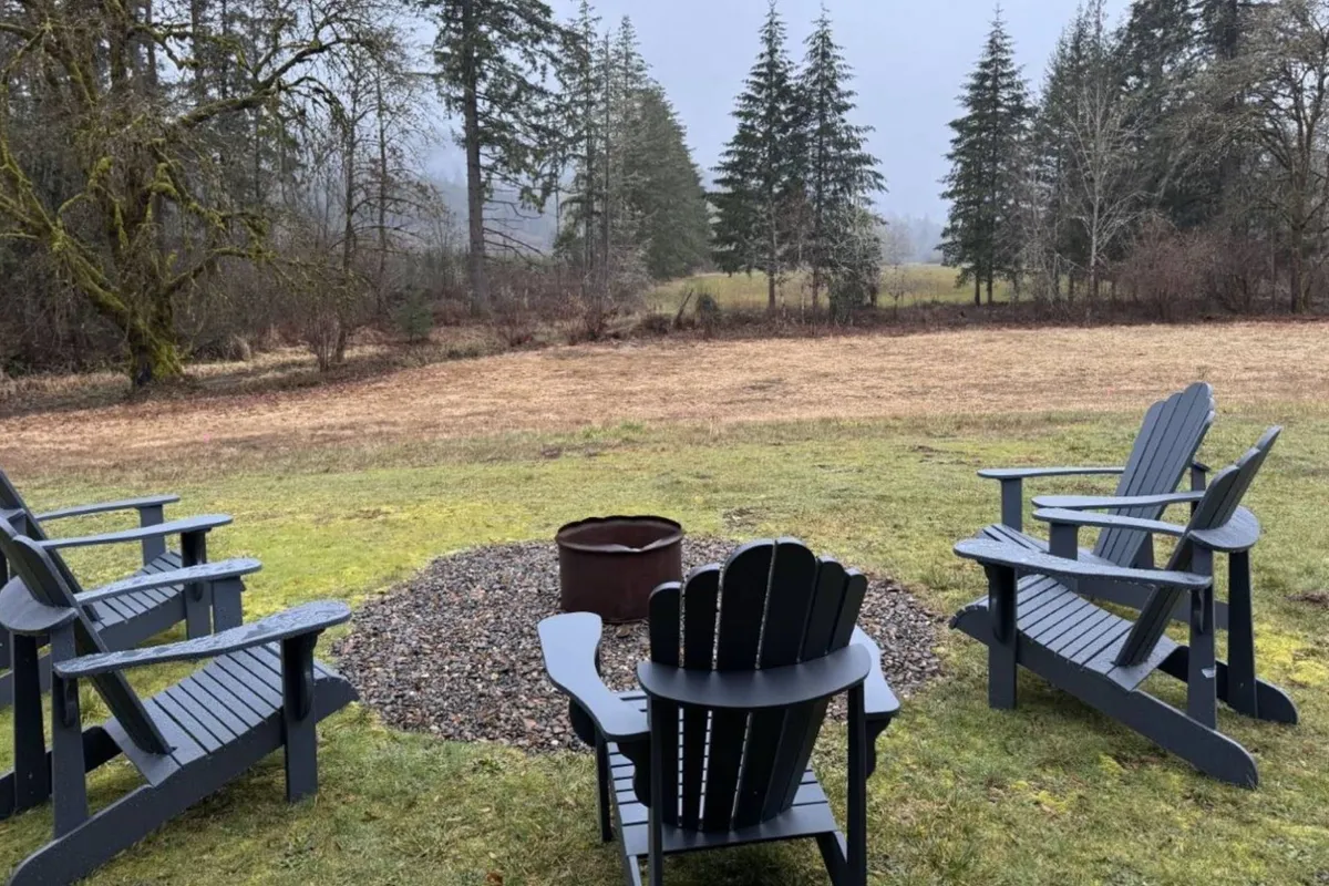 A tranquil scene with Adirondack chairs encircling a fire pit on a green lawn, bordered by trees and open fields. The setup is ideal for socializing, storytelling, and immersing in the peacefulness of rural living, especially under the stars or at sunrise.