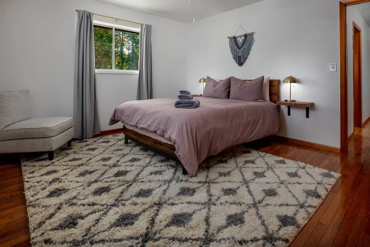 A calm and inviting bedroom featuring a comfortable bed, a soft rug, a window allowing in gentle daylight, an armchair, and tasteful wall decor that together evoke a cozy, restful ambiance.