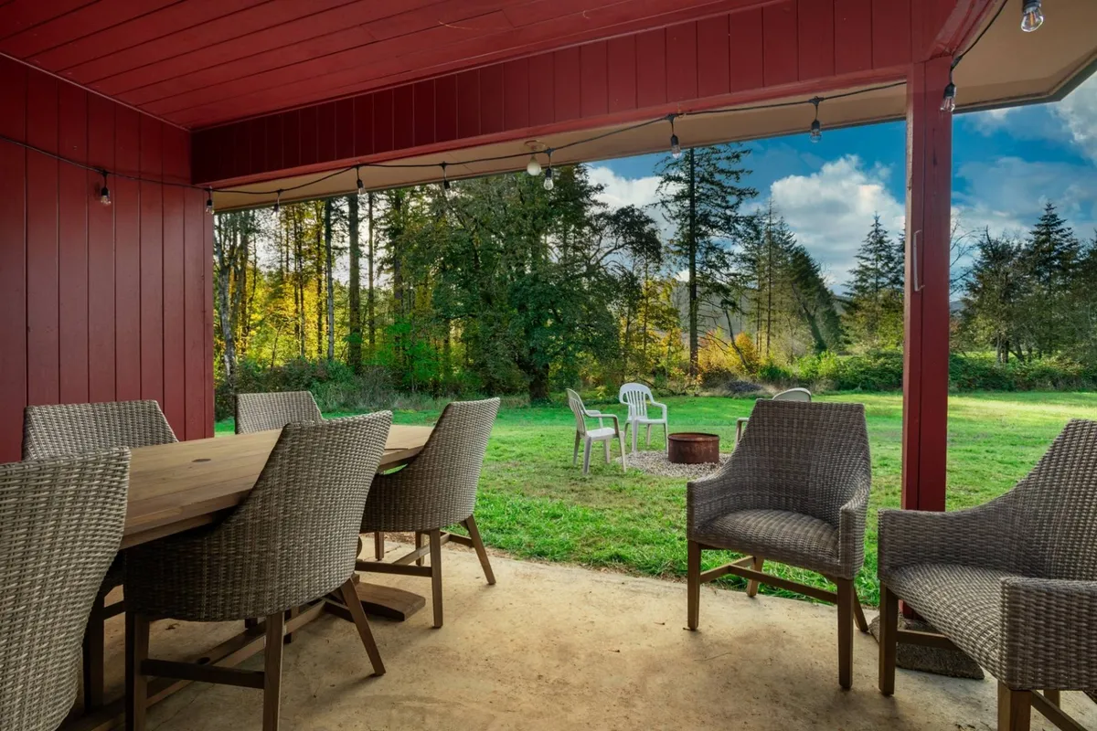 This exterior image features a spacious outdoor area with a dining table, lounge chairs arranged around a fire pit, and a lush green lawn bordered by mature trees. The setting invites gatherings, relaxation, and dining under the open sky, with nature as the backdrop.