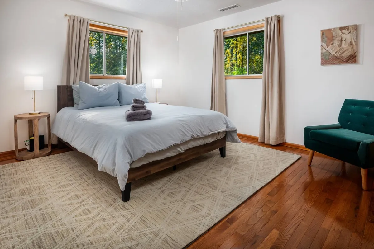 A serene queen bedroom with a plush bed, sunlit windows, a comfortable armchair, and matching nightstands. The soft light creates a peaceful atmosphere, making it an inviting retreat for relaxation or quiet reading.