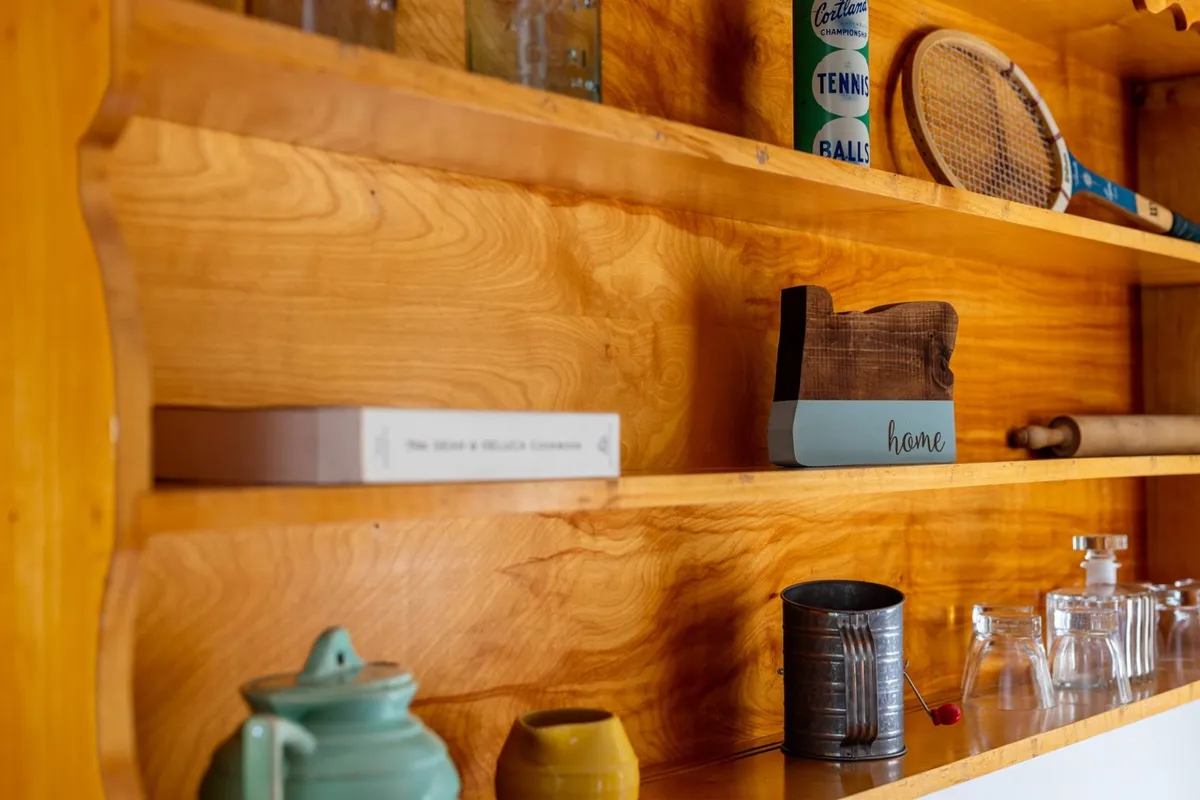 A cozy and welcoming shelf display with wooden shelving, a collection of glassware, a wooden block labeled 'home,' a tennis racket, and various decorative items. The assortment hints at an active lifestyle and a home base full of personality and charm.